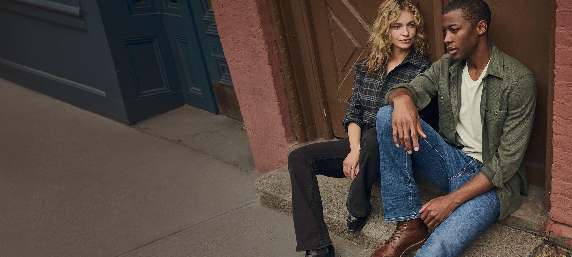 Couple sitting against a wall wearing fresh Lee bootcut denim. 