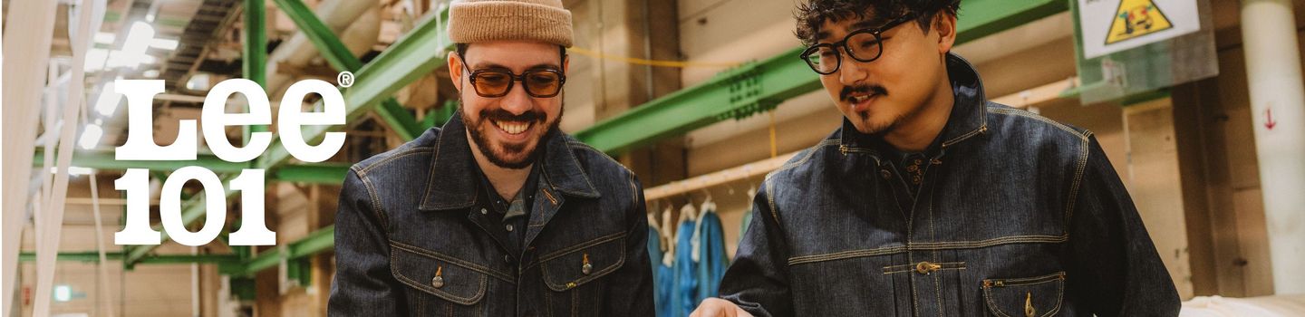 Due uomini con indosso giacche di jeans e occhiali da vista stanno ispezionando un grande rotolo di tessuto in un magazzino tessile. Uno ha la barba e sorride, mentre l'altro indica il tessuto. Il logo Lee 101 è ben visibile nell'angolo.