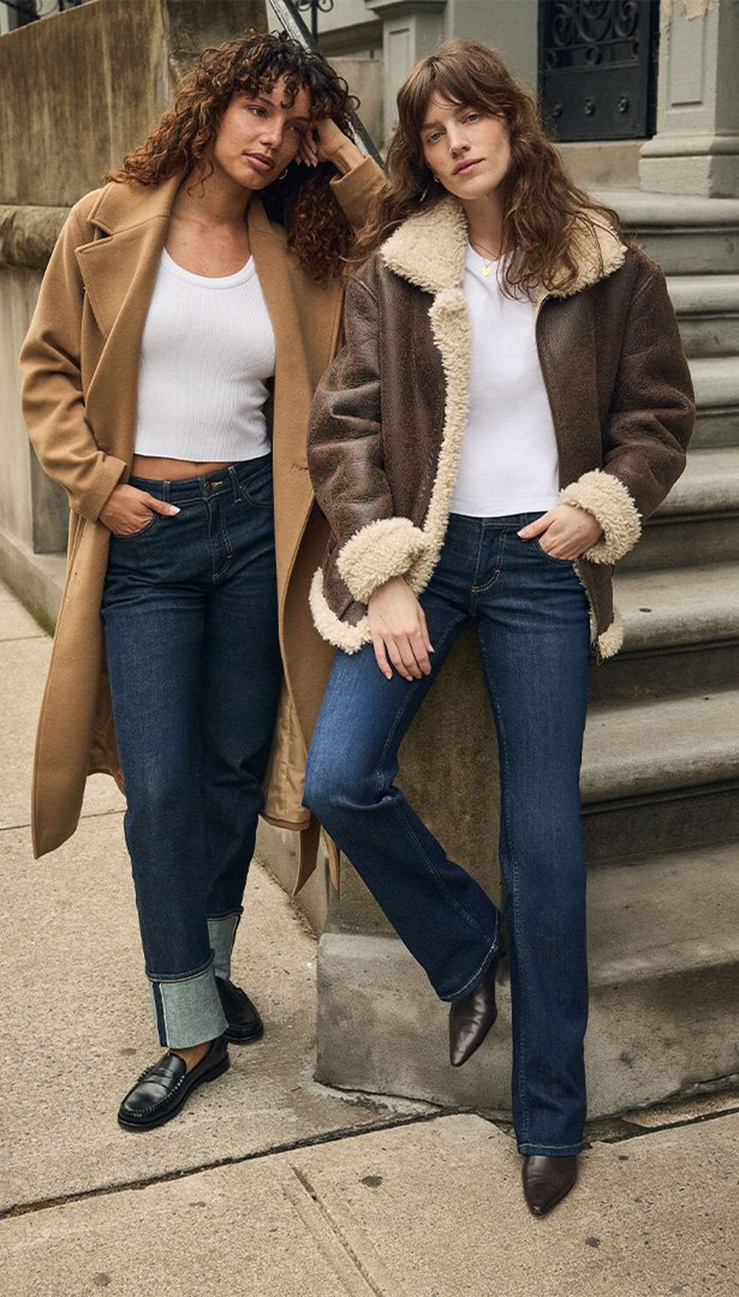 Dos mujeres con ropa de otoño posan en una acera: una lleva un abrigo cámel y la otra una chaqueta de zalea.