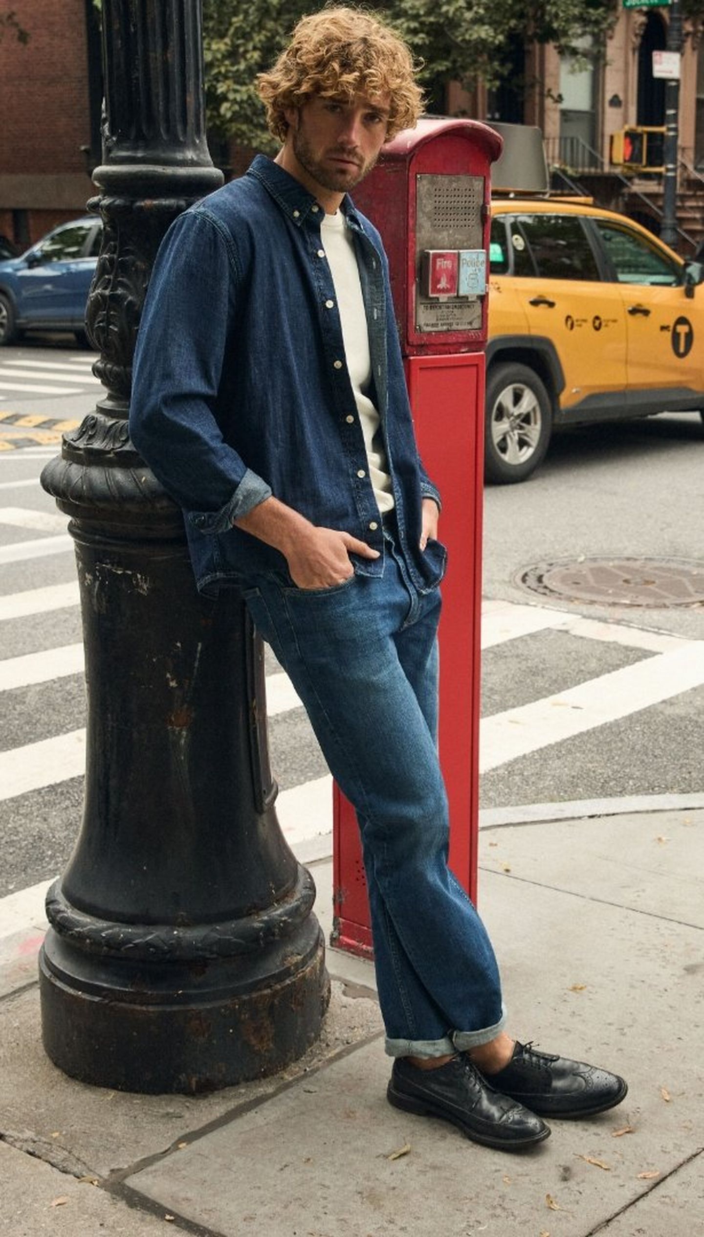 Man in denim shirt and jeans leans against a lamppost on a city street, with a yellow taxi in the background.